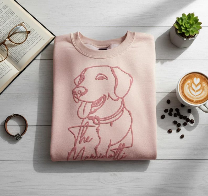 May include: A light pink sweatshirt with a dog graphic and the text "The Marsiliotti". The sweatshirt is next to a cup of coffee, a succulent plant, an open book, and a bracelet.