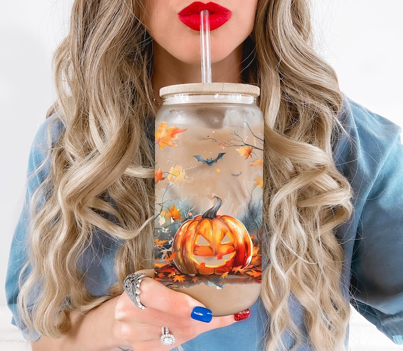 Puede incluir: Un vaso de vidrio transparente con un dise&ntilde;o de Halloween en negro, naranja y marr&oacute;n que presenta una calabaza y murci&eacute;lagos. El vaso tiene una tapa de bamb&uacute; y una pajita.
