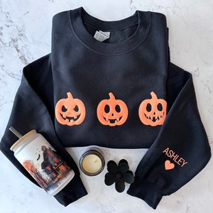 Peut inclure: Sweat-shirt noir avec trois visages de citrouille orange et le mot "ASHLEY" sur la manche. Un c&oelig;ur est imprim&eacute; &agrave; c&ocirc;t&eacute; du nom.