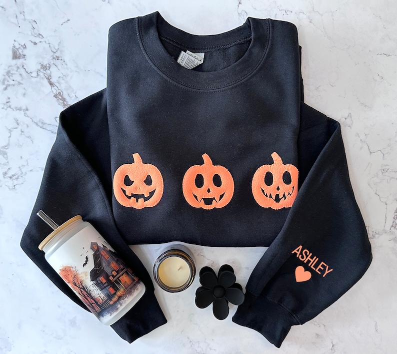 Peut inclure: Sweat-shirt noir avec trois visages de citrouille orange et le mot "ASHLEY" sur la manche. Un c&oelig;ur est imprim&eacute; &agrave; c&ocirc;t&eacute; du nom.