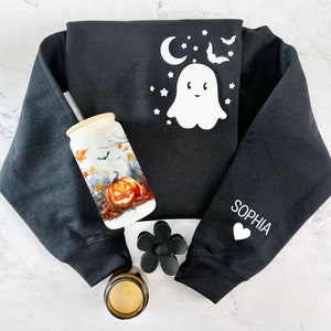 Puede incluir: Sudadera negra con un dise&ntilde;o de fantasma blanco y murci&eacute;lagos en la parte delantera y el nombre "Sophia" en la manga. La sudadera est&aacute; sobre una superficie de m&aacute;rmol blanco con un vaso de cer&aacute;mica blanco con un dise&ntilde;o de calabaza y una pajita, una flor negra y una vela.