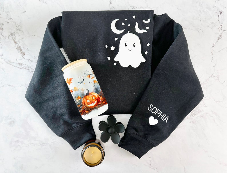 Puede incluir: Sudadera negra con un dise&ntilde;o de fantasma blanco y murci&eacute;lagos en la parte delantera y el nombre "Sophia" en la manga. La sudadera est&aacute; sobre una superficie de m&aacute;rmol blanco con un vaso de cer&aacute;mica blanco con un dise&ntilde;o de calabaza y una pajita, una flor negra y una vela.