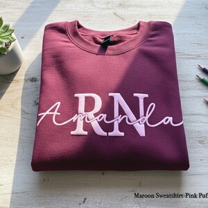 以下が含まれることがあります： ライトピンクの筆記体で「RN Amanda」と書かれた、マルーン色のスウェットシャツ。スウェットシャツは明るい色の木製の表面にきれいに折りたたまれています。小さな鉢植えの多肉植物と色鉛筆が背景にあります。