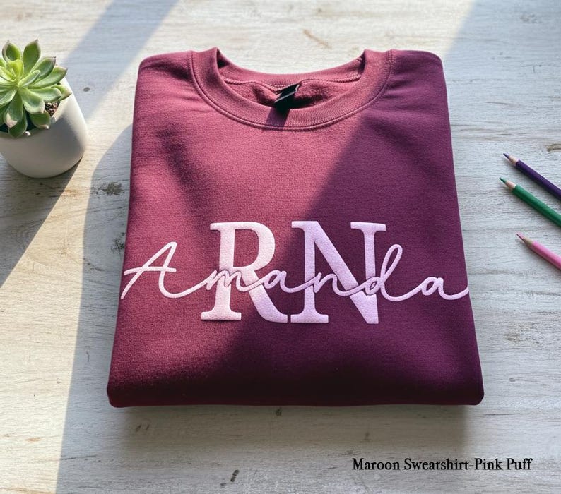 以下が含まれることがあります： ライトピンクの筆記体で「RN Amanda」と書かれた、マルーン色のスウェットシャツ。スウェットシャツは明るい色の木製の表面にきれいに折りたたまれています。小さな鉢植えの多肉植物と色鉛筆が背景にあります。