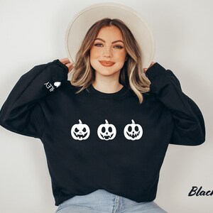 Peut inclure: Sweat-shirt noir avec un motif blanc de trois citrouilles souriantes. Le texte "HEY" est sur la manche gauche.