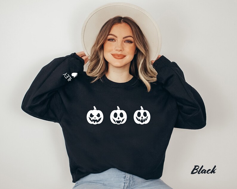 Peut inclure: Sweat-shirt noir avec un motif blanc de trois citrouilles souriantes. Le texte "HEY" est sur la manche gauche.