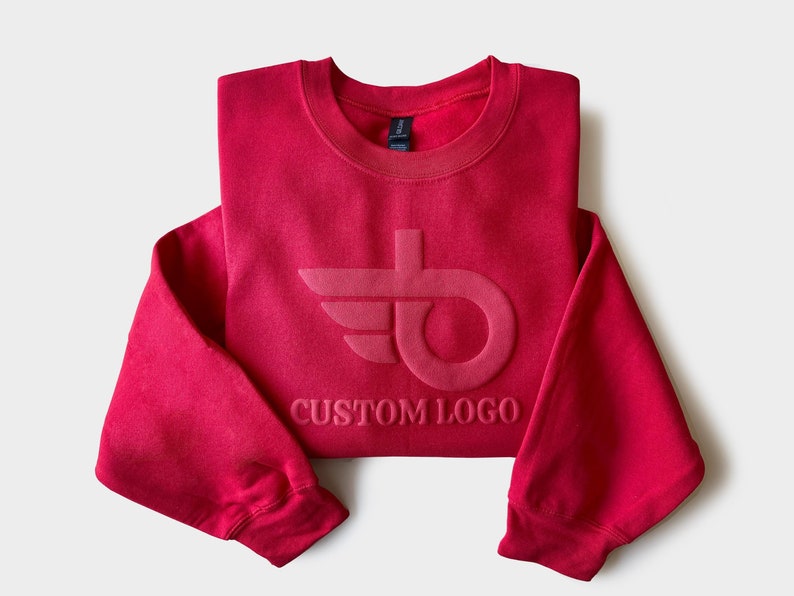 May include: A folded red sweatshirt with a crew neck and long sleeves. The sweatshirt features a tonal custom logo design on the front. The text "CUSTOM LOGO" is below the logo. The garment is made of a soft material.