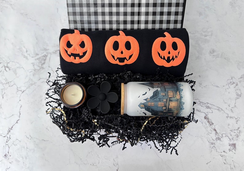 Peut inclure: Une bo&icirc;te cadeau sur le th&egrave;me d'Halloween avec un sac &agrave; carreaux noir et blanc, un tissu noir avec trois citrouilles orange, une bougie noire et blanche, un moule en silicone noir en forme de fleur et un gobelet blanc avec un motif de maison hant&eacute;e.