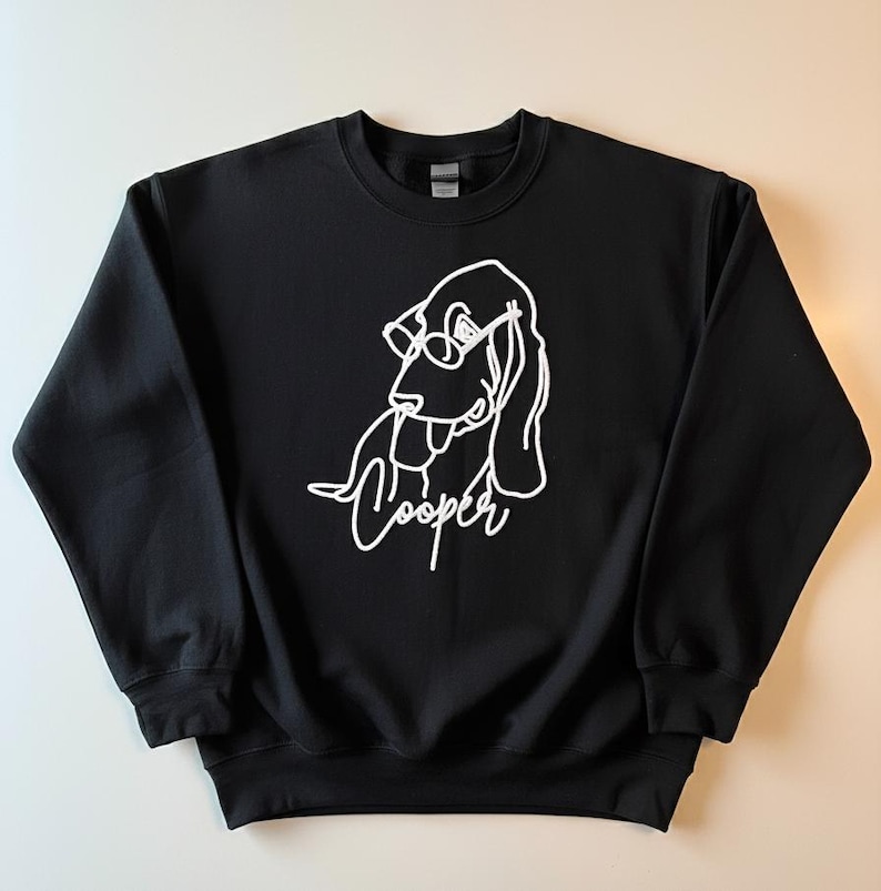 May include: Black crewneck sweatshirt featuring a white line drawing of a dog wearing glasses. The dog's name, "Cooper," is written in cursive below the drawing. The sweatshirt is made of a soft material.
