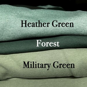 May include: A stack of three folded sweatshirts in shades of green. The top sweatshirt is heather green, the middle is forest green, and the bottom is military green. Text labels each color.