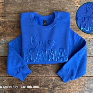 May include: Royal blue sweatshirt with the words "Boys Mama" in metallic blue lettering. The sweatshirt has a crew neck and long sleeves. A circular graphic with the same text is in the upper right corner.