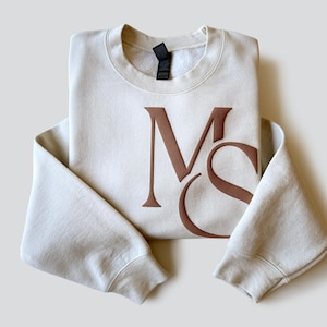 May include: Cream-colored sweatshirt with the brown letters "MS" printed on the front. The sweatshirt is folded neatly, with the sleeves extending outward. The fabric appears soft and comfortable, suitable for casual wear.