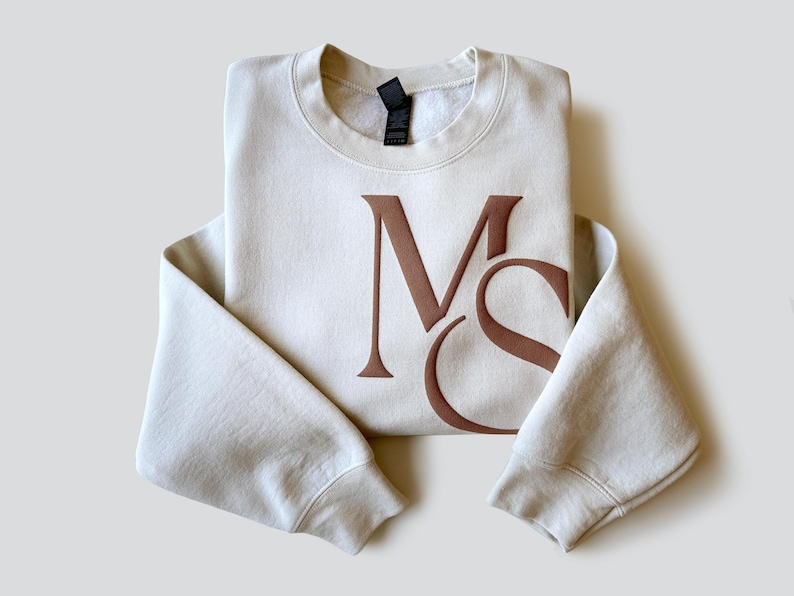 May include: Cream-colored sweatshirt with the brown letters "MS" printed on the front. The sweatshirt is folded neatly, with the sleeves extending outward. The fabric appears soft and comfortable, suitable for casual wear.
