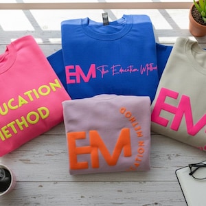 May include: Four sweatshirts in pink, blue, lavender, and beige, each with a different color of text. The pink sweatshirt has the words "The EDUCATION METHOD" in yellow. The blue sweatshirt has "EM The Education Method" in pink.
