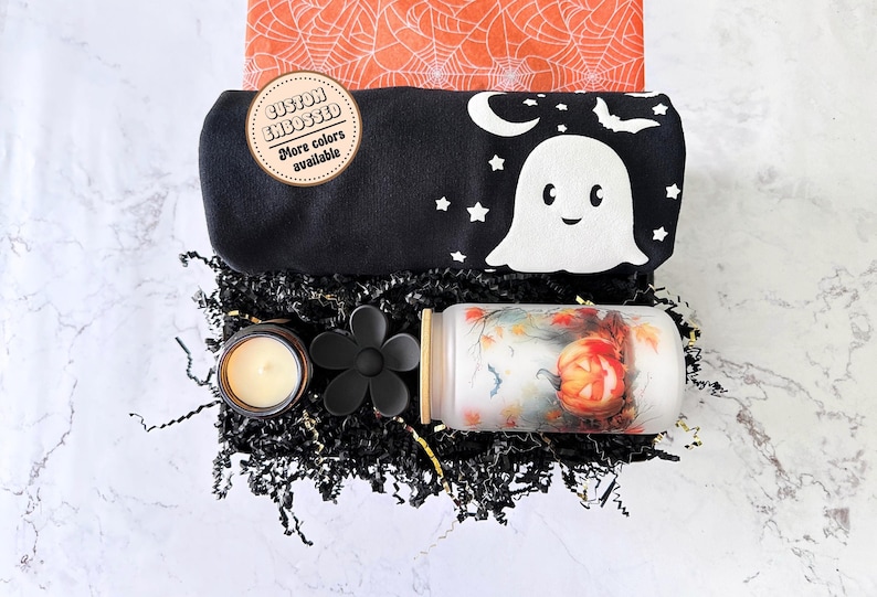 Puede incluir: Una caja de regalo de Halloween con una tela negra y blanca con un dise&ntilde;o de fantasma, un portavelas negro con forma de flor, una vela de vidrio marr&oacute;n y una vela blanca con un dise&ntilde;o de calabaza.