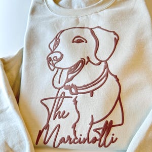 May include: Cream-colored sweatshirt featuring a brown outline of a dog's face and the text "The Marcintotti." The dog has its tongue out. The sweatshirt has a crew neck and long sleeves.