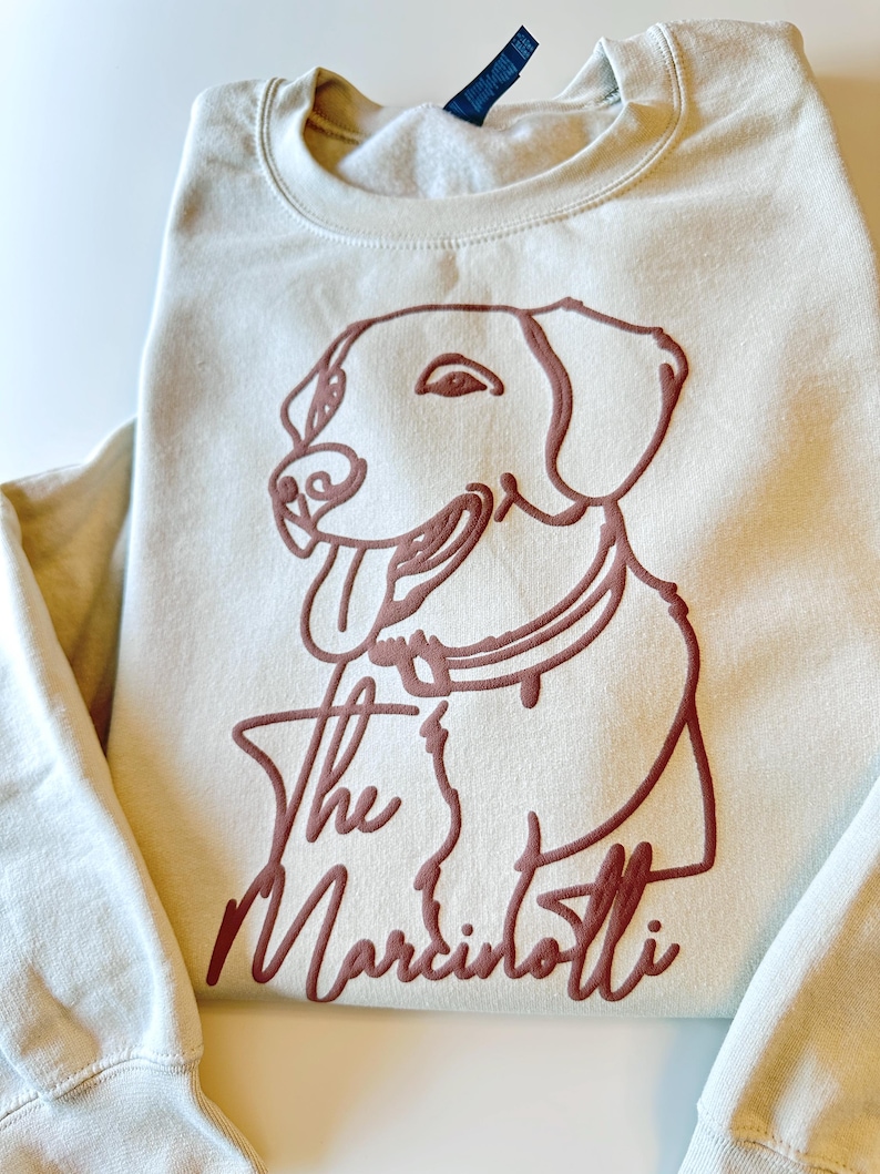 May include: Cream-colored sweatshirt featuring a brown outline of a dog's face and the text "The Marcintotti." The dog has its tongue out. The sweatshirt has a crew neck and long sleeves.