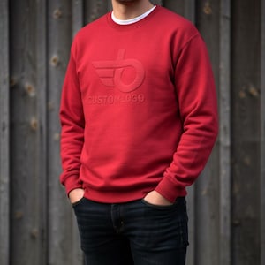May include: A red sweatshirt with a crew neck and long sleeves. The sweatshirt features a tonal embossed logo with the text "CUSTOM LOGO". The person is wearing dark blue jeans and a white undershirt.