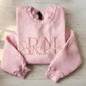 以下が含まれることがあります： 「Sophia」の名前の上に「RN」の刺繍が入った、明るいピンク色のスウェットシャツ。 スウェットシャツは明るい木製の表面にきれいに折りたたまれています。 白い鉢に入った小さな多肉植物が背景に見えます。