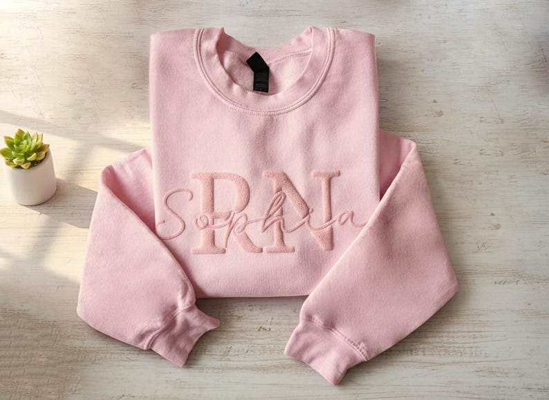 以下が含まれることがあります： 「Sophia」の名前の上に「RN」の刺繍が入った、明るいピンク色のスウェットシャツ。 スウェットシャツは明るい木製の表面にきれいに折りたたまれています。 白い鉢に入った小さな多肉植物が背景に見えます。
