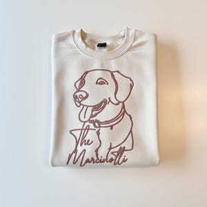 May include: Cream-colored sweatshirt with a maroon outline of a dog and the text "The Marciolotti." The dog is smiling with its tongue out. The sweatshirt is folded neatly.