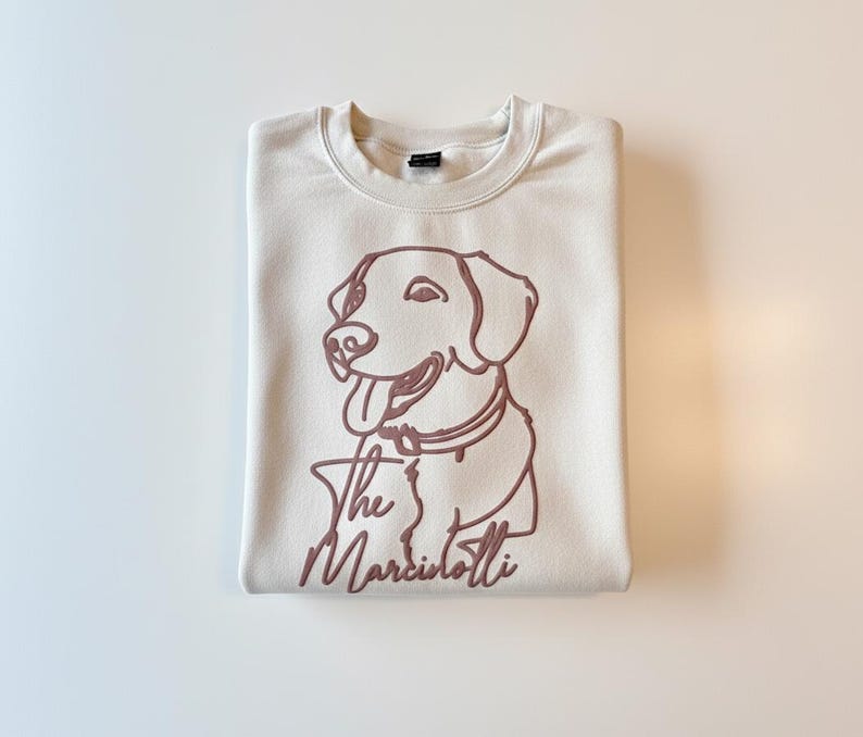 May include: Cream-colored sweatshirt with a maroon outline of a dog and the text "The Marciolotti." The dog is smiling with its tongue out. The sweatshirt is folded neatly.