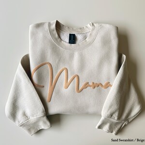 May include: A sand-colored sweatshirt with the word "Mama" embroidered in a light brown script across the front. The sweatshirt has a crew neck and long sleeves. The text "Sand Sweatshirt / Beige Puff" is at the bottom.