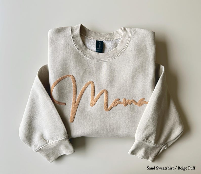 May include: A sand-colored sweatshirt with the word "Mama" embroidered in a light brown script across the front. The sweatshirt has a crew neck and long sleeves. The text "Sand Sweatshirt / Beige Puff" is at the bottom.