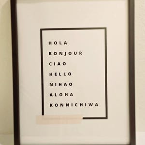 May include: A black framed print with a white background and a black rectangle in the center. The rectangle contains the words "Hola", "Bonjour", "Ciao", "Hello", "Nihao", "Aloha", and "Konnichiwa" in black text.
