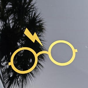 May include: Yellow vinyl decal of a lightning bolt and round glasses, a symbol from a popular fantasy series. The decal is affixed to a black surface, with a blurred tree reflection in the background.