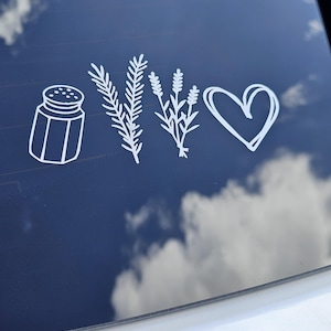 May include: White vinyl decal featuring a salt shaker, sprigs of rosemary and lavender, and a heart. The decal is designed to be applied to a car window or similar surface. The background is a dark blue with clouds.