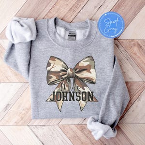 K&ouml;nnte beinhalten: Sport Grey Sweatshirt mit einer gro&szlig;en Schleife im Camouflage-Muster. Die Schleife ist mittig auf der Vorderseite platziert und darunter steht der Text "Mrs. JOHNSON" in einer fetten, blockartigen Schrift. Das Sweatshirt hat einen Rundhalsausschnitt.