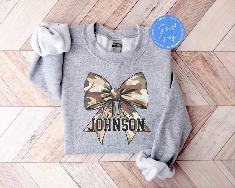 K&ouml;nnte beinhalten: Sport Grey Sweatshirt mit einer gro&szlig;en Schleife im Camouflage-Muster. Die Schleife ist mittig auf der Vorderseite platziert und darunter steht der Text "Mrs. JOHNSON" in einer fetten, blockartigen Schrift. Das Sweatshirt hat einen Rundhalsausschnitt.