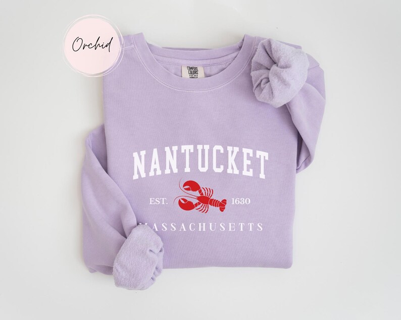 May include: A light purple sweatshirt with the word "NANTUCKET" in white block letters, a red lobster graphic, and the text "EST. 1630" and "MASSACHUSETTS" in white. The word "Orchid" is in a pink circle in the upper left corner.
