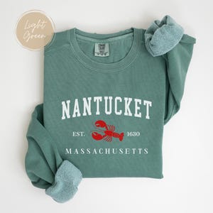 May include: A light green sweatshirt with the word "NANTUCKET" in white block letters. Below the word is a red lobster graphic and the words "EST. 1630" and "MASSACHUSETTS" in white. The sweatshirt has a crew neck and long sleeves.