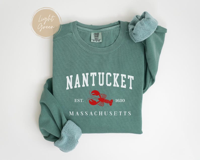 May include: A light green sweatshirt with the word "NANTUCKET" in white block letters. Below the word is a red lobster graphic and the words "EST. 1630" and "MASSACHUSETTS" in white. The sweatshirt has a crew neck and long sleeves.