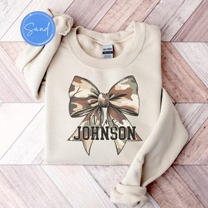 K&ouml;nnte beinhalten: Beiges Sweatshirt mit gro&szlig;em Schleifendesign im Camouflage-Muster. Der Text "Mrs. JOHNSON" ist unterhalb der Schleife in fetter Schrift gedruckt. Das Sweatshirt ist gefaltet, die &Auml;rmel sind sichtbar.