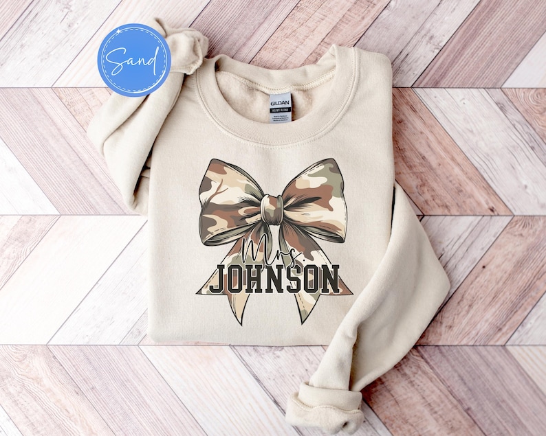 K&ouml;nnte beinhalten: Beiges Sweatshirt mit gro&szlig;em Schleifendesign im Camouflage-Muster. Der Text "Mrs. JOHNSON" ist unterhalb der Schleife in fetter Schrift gedruckt. Das Sweatshirt ist gefaltet, die &Auml;rmel sind sichtbar.