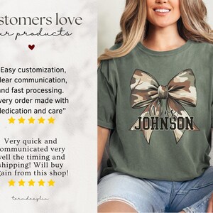 K&ouml;nnte beinhalten: Olivgr&uuml;nes T-Shirt mit einem gro&szlig;en Schleifendesign im Camouflage-Muster und dem Namen "Mrs. JOHNSON". Das T-Shirt hat eine lockere Passform. Das Bild enth&auml;lt auch Kundenreferenzen. Der Text auf dem T-Shirt ist in einer Serifenschrift.