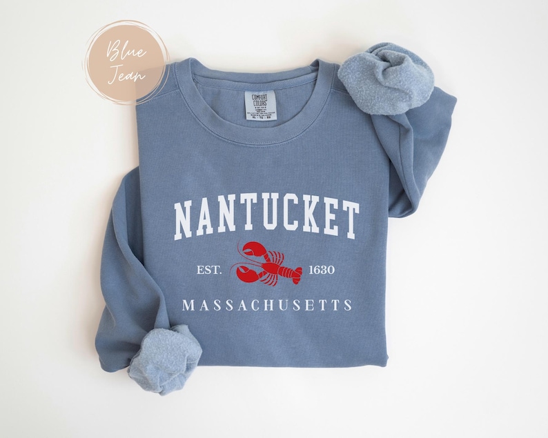 May include: A blue sweatshirt with the word "NANTUCKET" in white block letters. Below the word is a red lobster graphic and the text "EST. 1630" and "MASSACHUSETTS". The sweatshirt has long sleeves and a crew neck.