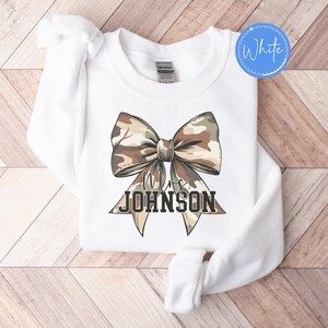 K&ouml;nnte beinhalten: Wei&szlig;es Sweatshirt mit Rundhalsausschnitt und einer gro&szlig;en Tarnschleife. Das Schleifendesign hat braune, beige und gr&uuml;ne Farbt&ouml;ne. Unter der Schleife steht der Text "Mrs. JOHNSON" in schwarzer, fetter Schrift. Ein wei&szlig;er Kreis mit dem Wort "White" befindet sich in der oberen rechten Ecke.