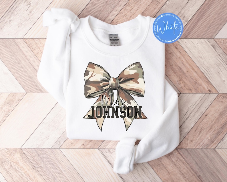 K&ouml;nnte beinhalten: Wei&szlig;es Sweatshirt mit Rundhalsausschnitt und einer gro&szlig;en Tarnschleife. Das Schleifendesign hat braune, beige und gr&uuml;ne Farbt&ouml;ne. Unter der Schleife steht der Text "Mrs. JOHNSON" in schwarzer, fetter Schrift. Ein wei&szlig;er Kreis mit dem Wort "White" befindet sich in der oberen rechten Ecke.