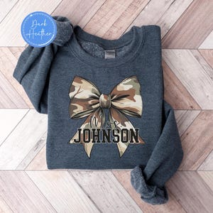 K&ouml;nnte beinhalten: Dunkelgraues Sweatshirt mit einer gro&szlig;en Camouflage-Schleife. Unter der Schleife steht der Text "Mrs. JOHNSON". Das Sweatshirt hat einen Rundhalsausschnitt und lange &Auml;rmel. Das Kleidungsst&uuml;ck hat eine dunkel melierte Farbe.