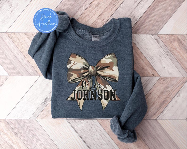 K&ouml;nnte beinhalten: Dunkelgraues Sweatshirt mit einer gro&szlig;en Camouflage-Schleife. Unter der Schleife steht der Text "Mrs. JOHNSON". Das Sweatshirt hat einen Rundhalsausschnitt und lange &Auml;rmel. Das Kleidungsst&uuml;ck hat eine dunkel melierte Farbe.