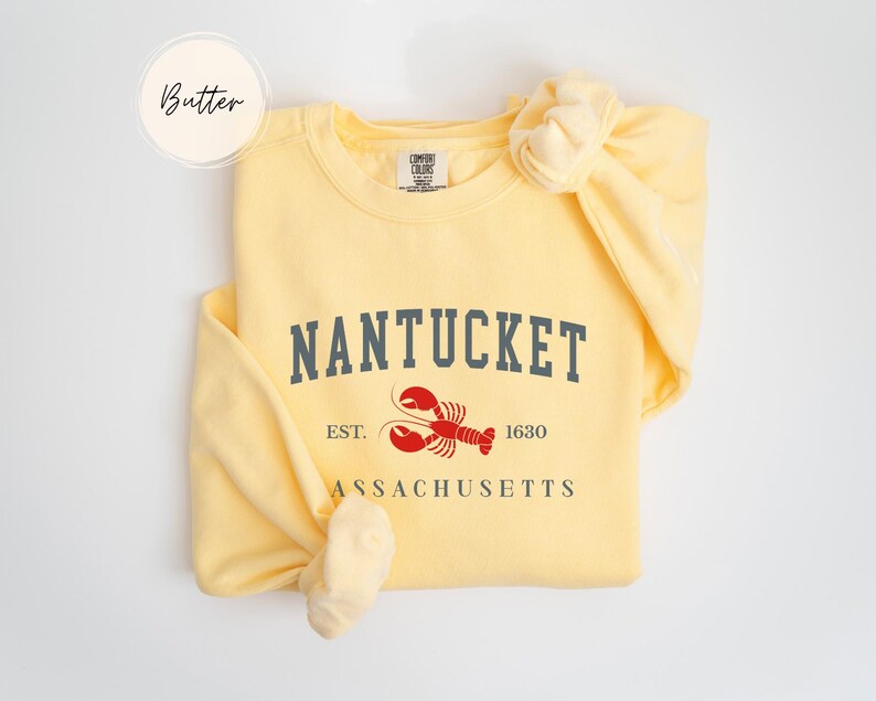 May include: A light yellow sweatshirt with the word "Nantucket" in gray block letters, a red lobster graphic, and the text "Est. 1630 Massachusetts". The word "Butter" is in a circle in the upper left.