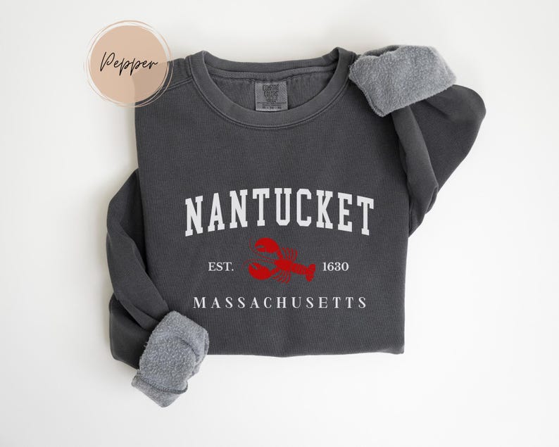 May include: Dark gray sweatshirt with the word "NANTUCKET" in white block letters, a red lobster graphic, and the text "EST. 1630 MASSACHUSETTS". The sweatshirt has a crew neck and gray fleece cuffs.