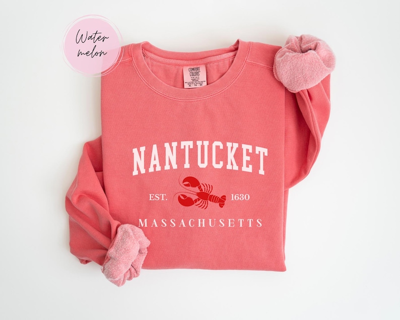 May include: A coral-colored sweatshirt with the word "NANTUCKET" in white block letters. Below the word is a red lobster graphic and the text "EST. 1630" and "MASSACHUSETTS". A pink scrunchie is on the sleeve. The word "Watermelon" is in a pink circle.