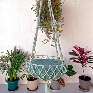 May include: A light green macrame cat hammock with a leopard print cushion. The hammock is hanging from the ceiling and there are plants in the background.