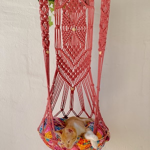May include: A pink macrame cat hammock with a colorful patterned cushion. A cat is sleeping in the hammock.