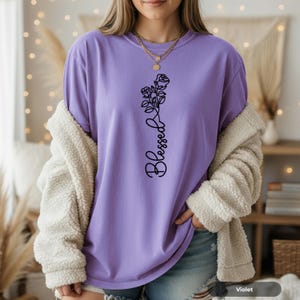 Puede incluir: Una camiseta de color violeta con un diseño floral negro y la palabra "Blessed" impresa verticalmente. La camiseta es de corte holgado, y la modelo lleva una chaqueta color crema y vaqueros azules.